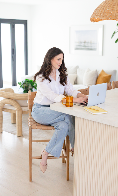 A woman with long brown hair sits on a wooden stool at a kitchen island, working on a laptop. She is wearing a white button-down shirt and light-wash jeans, with a mug and notebook beside her.