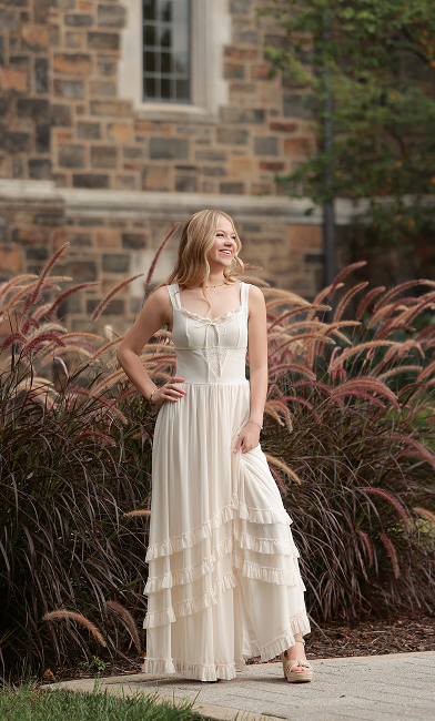 A young woman with blonde hair stands smiling in a long, cream-colored dress with ruffled tiers, posing in front of tall ornamental grass and a stone building. She is holding up the hem of her dress and wearing platform sandals.