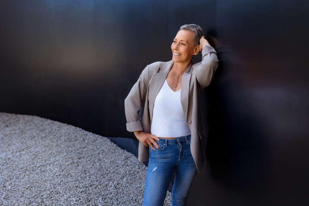 Smiling woman with short gray hair, wearing a beige blazer, white top, and jeans, leans against a dark wall