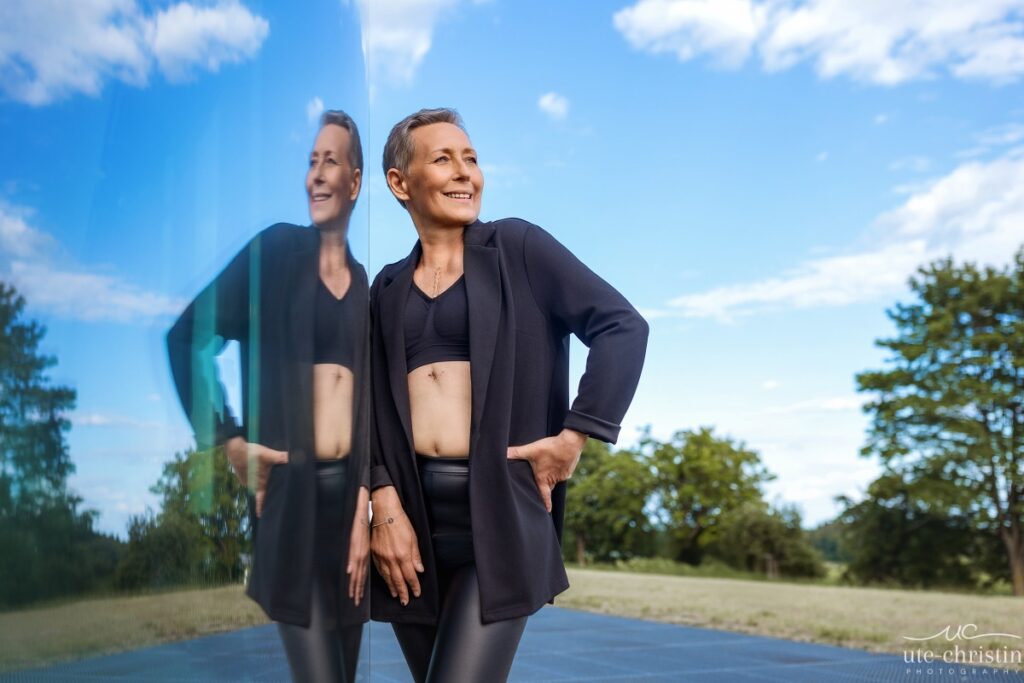 Smiling woman with short gray hair, wearing black top, jacket, and pants, stands by reflective surface outdoors