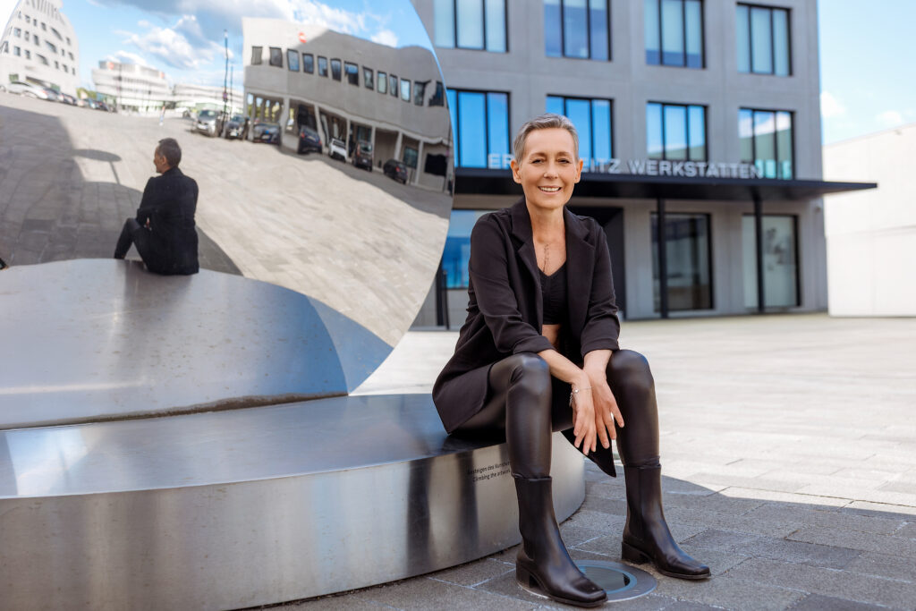 Smiling woman in black sits on a metal sculpture, modern building in background