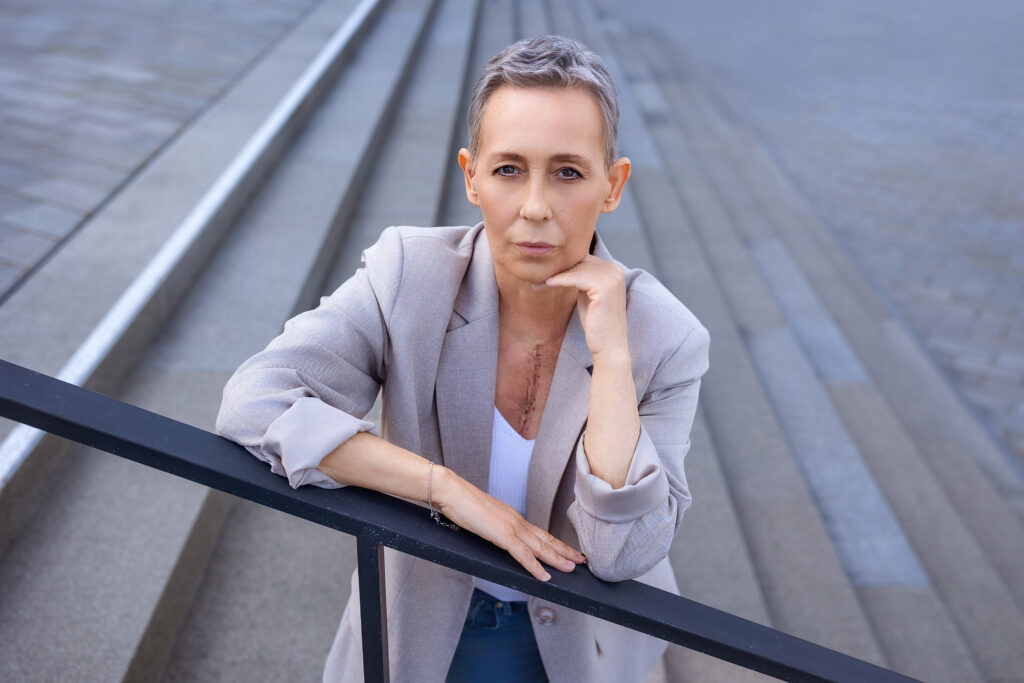 Serious mature woman with short gray hair, wearing a beige blazer, leans on a black railing on a staircase