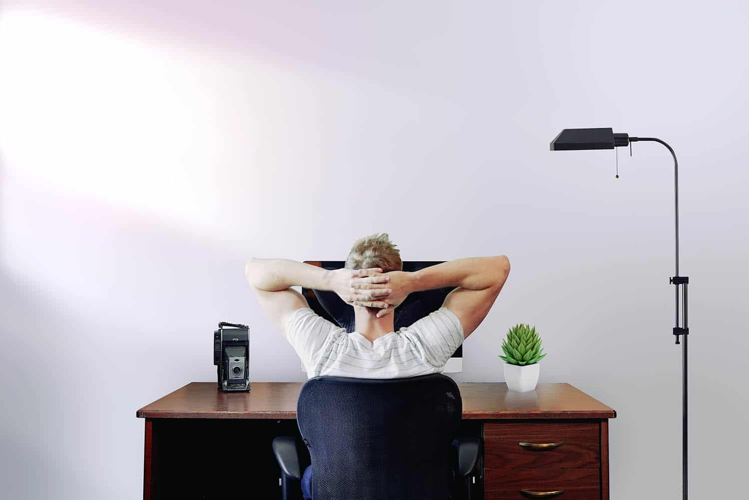 A white man in a white t-shirt sits at his desk in front of a white wall. He's looking at his computer monitor with his hands folded behind his head. He's learning to batch edit in Lightroom.