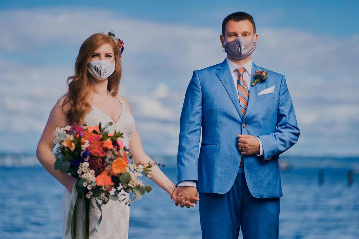 A bride and groom hold hands in front of the ocean with COVID-19 masks on their faces