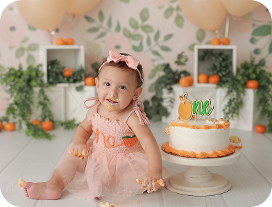 A baby girl in a peach-colored dress with "one" embroidered on the front sits next to a white cake with orange frosting and a "one" topper, surrounded by a floral backdrop, greenery, and oranges. The baby has cake on her face and hands, indicating a "smash cake" first birthday celebration.