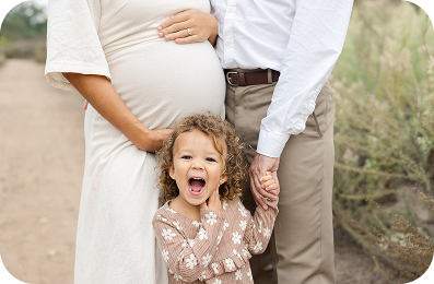 A pregnant woman in a cream dress holds her belly while standing next to a man in a white shirt and khaki pants, and their young daughter with curly hair and a floral shirt stands in front, smiling with her mouth open and holding their hands. The family is outdoors with a dirt path and foliage in the background.