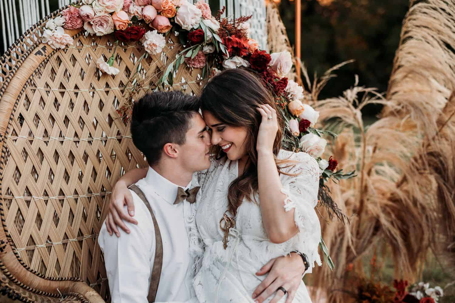 A boho bride sits in her husband's lap in a huge wicker chair
