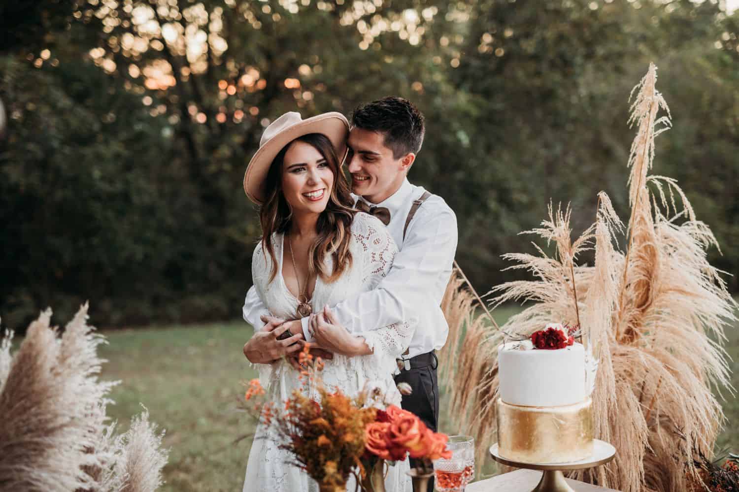 A boho bride and groom hug each other in front of their cake table