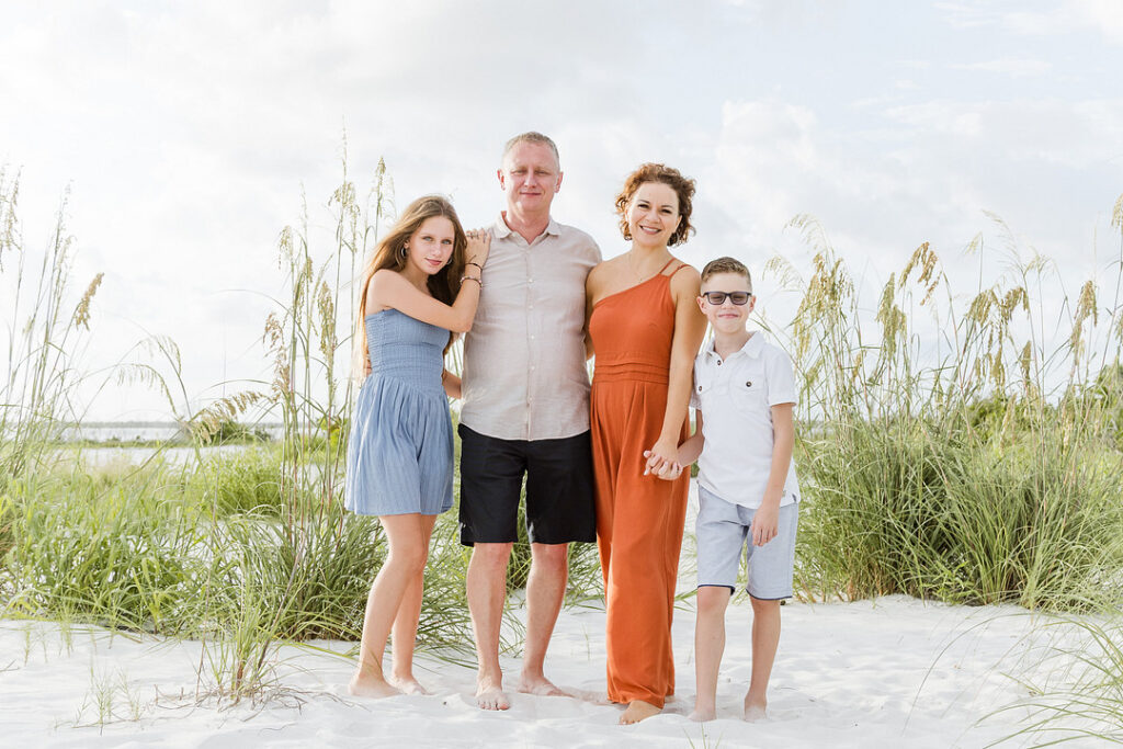 A family of four poses barefoot on a white sand beach, with tall grasses behind them and a bright sky overhead. The mother wears an orange jumpsuit, the father a tan shirt and black shorts, the daughter a blue dress, and the son a white polo and blue shorts.
