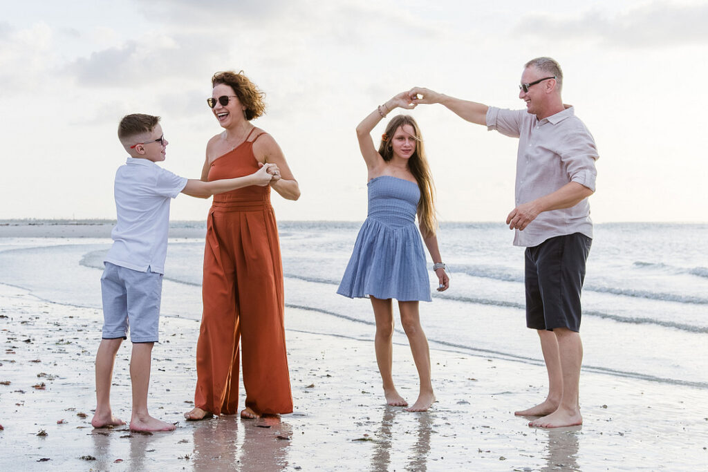 A family of four is dancing barefoot on a beach, with the mother in an orange jumpsuit dancing with her son, and the father in a light shirt and dark shorts dancing with his daughter. The scene is set against a backdrop of a calm ocean and a cloudy sky.