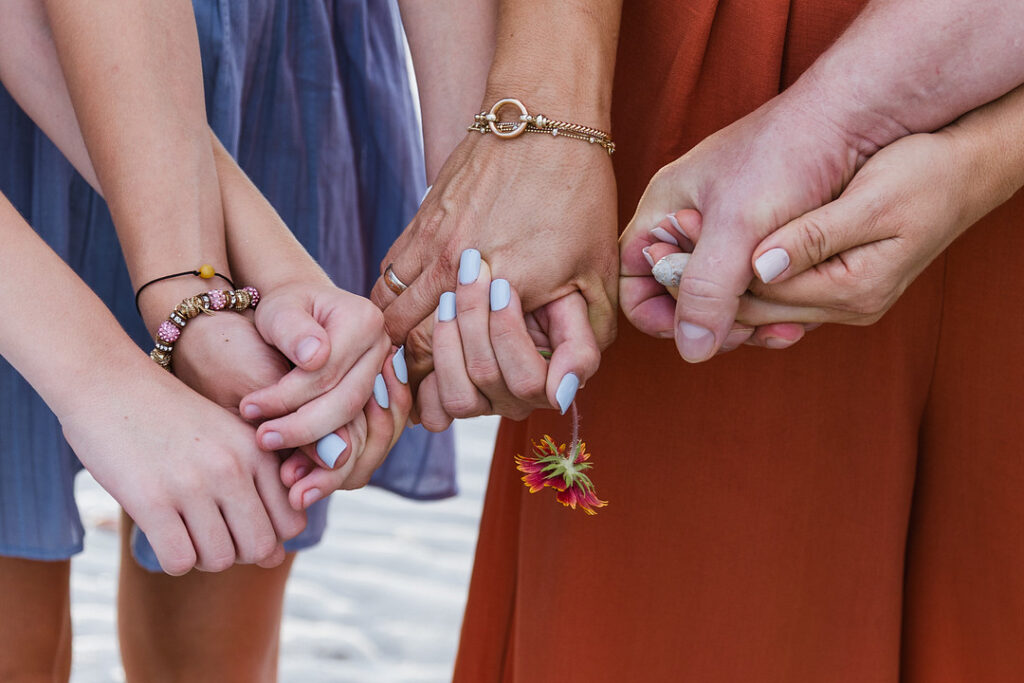 Three people hold hands, with the person in the middle wearing a gold bracelet and a wedding ring, and holding a small flower. The person on the right holds a small rock.