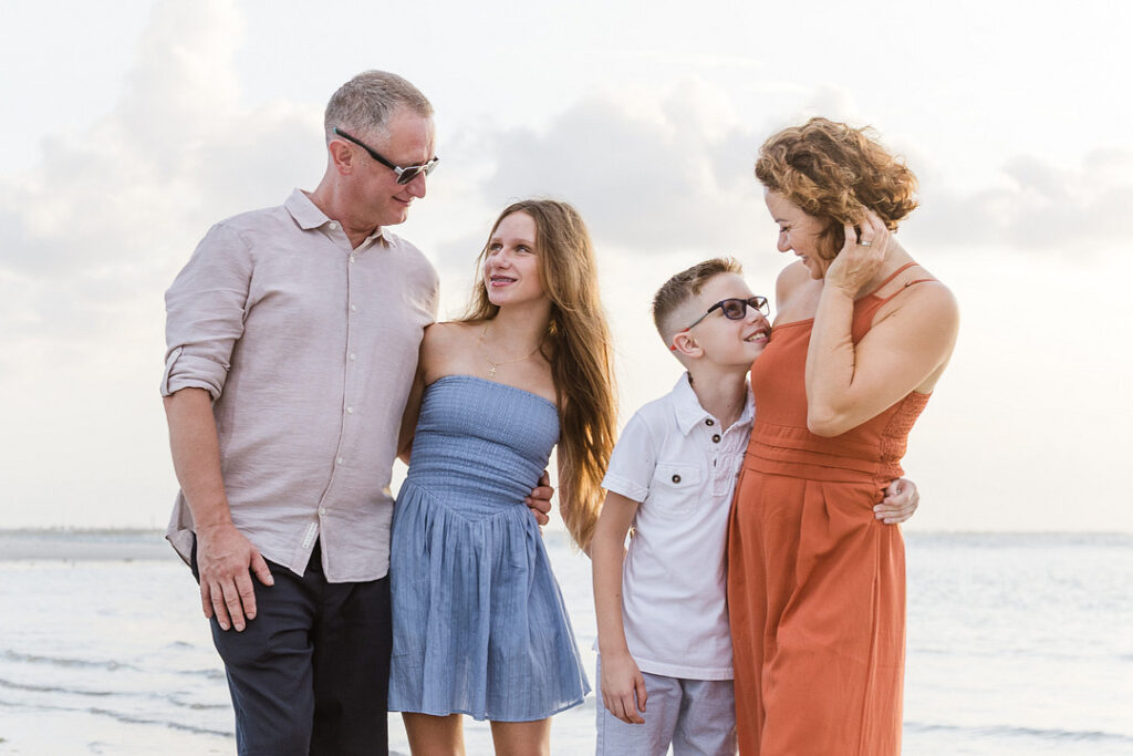 A family of four poses on a beach, with the parents wearing sunglasses and embracing their two children, a teenage girl in a blue dress and a younger boy in a white shirt. The family is smiling and looking at each other, with the ocean and a bright sky in the background.