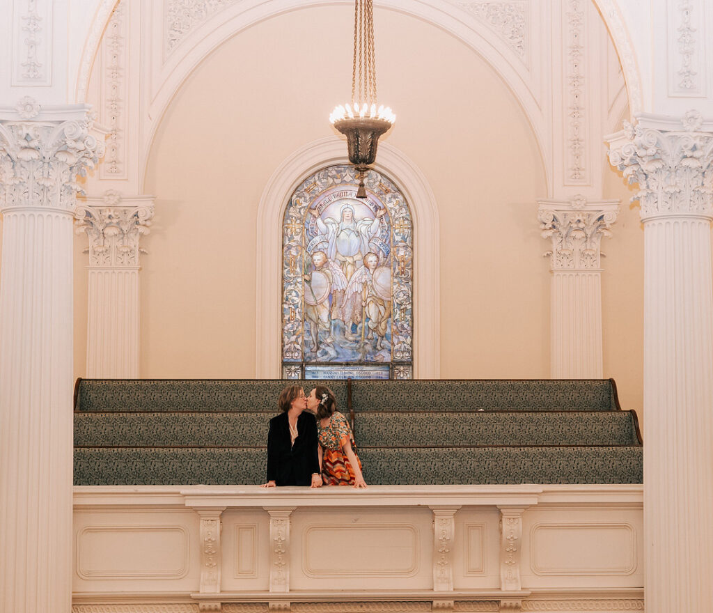Two women are kissing in a church, standing on a white balcony in front of green tiered seating, with a stained glass window and chandelier above them. The church features ornate white columns and arches.