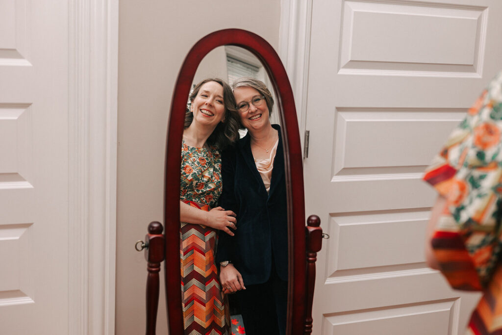 Two smiling women are reflected in a standing, dark wood-framed mirror; the woman on the left wears a floral top and chevron-patterned skirt, while the woman on the right wears a navy velvet suit.