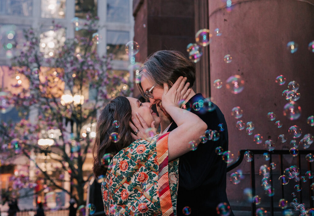 Two women embrace and kiss, surrounded by floating bubbles, with one woman wearing a floral patterned top and the other in a black jacket. A flowering tree and city buildings are visible in the background.