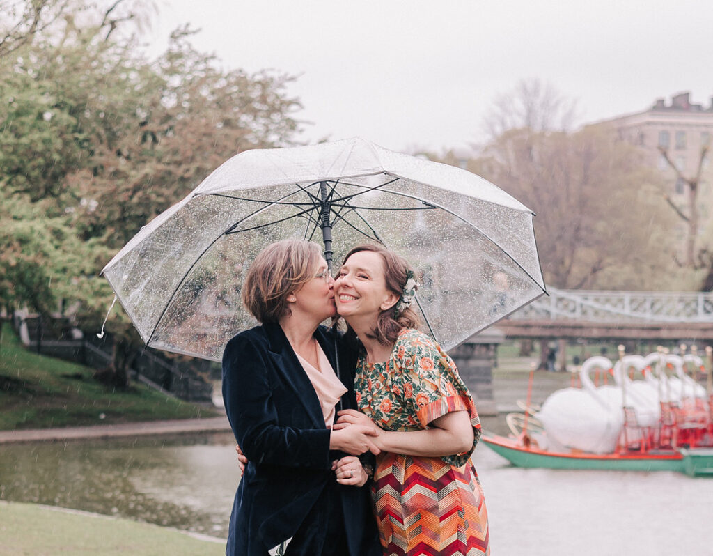 In the rain, two women stand under a clear umbrella, one kissing the other's cheek. They are in a park with a pond, and swan boats are visible in the background.