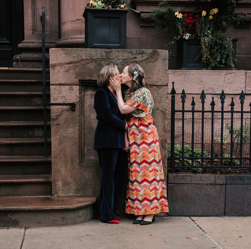 Two women, one in a navy velvet suit and red shoes and the other in a floral top and chevron-patterned skirt with green shoes, kiss in front of a brownstone building with flower boxes. The woman in the skirt has flowers in her hair and is holding the other woman's face.