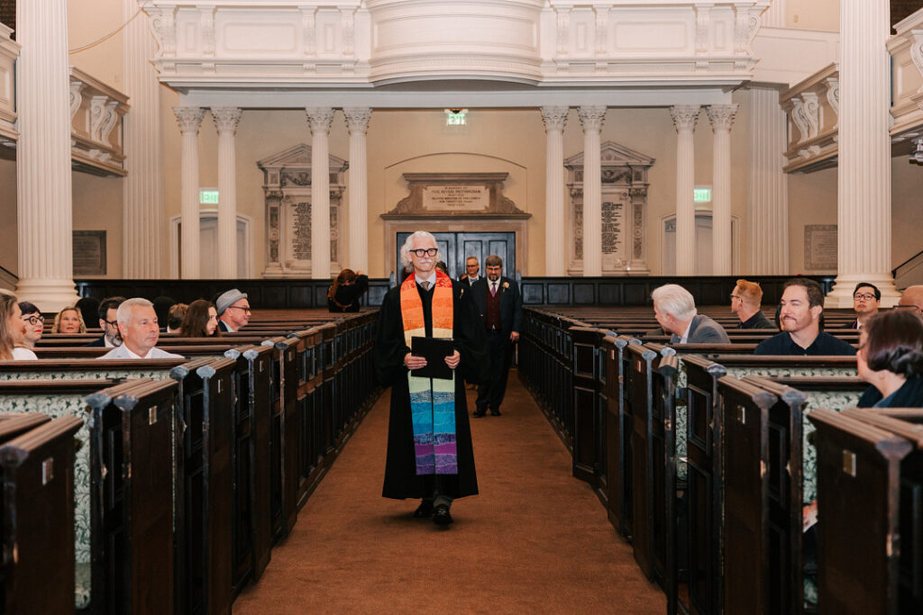 In a grand church interior, a minister in a black robe and colorful stole walks down the aisle, holding a black book, as wedding guests sit in the pews on either side. Two men in suits follow behind the minister, walking towards the camera.