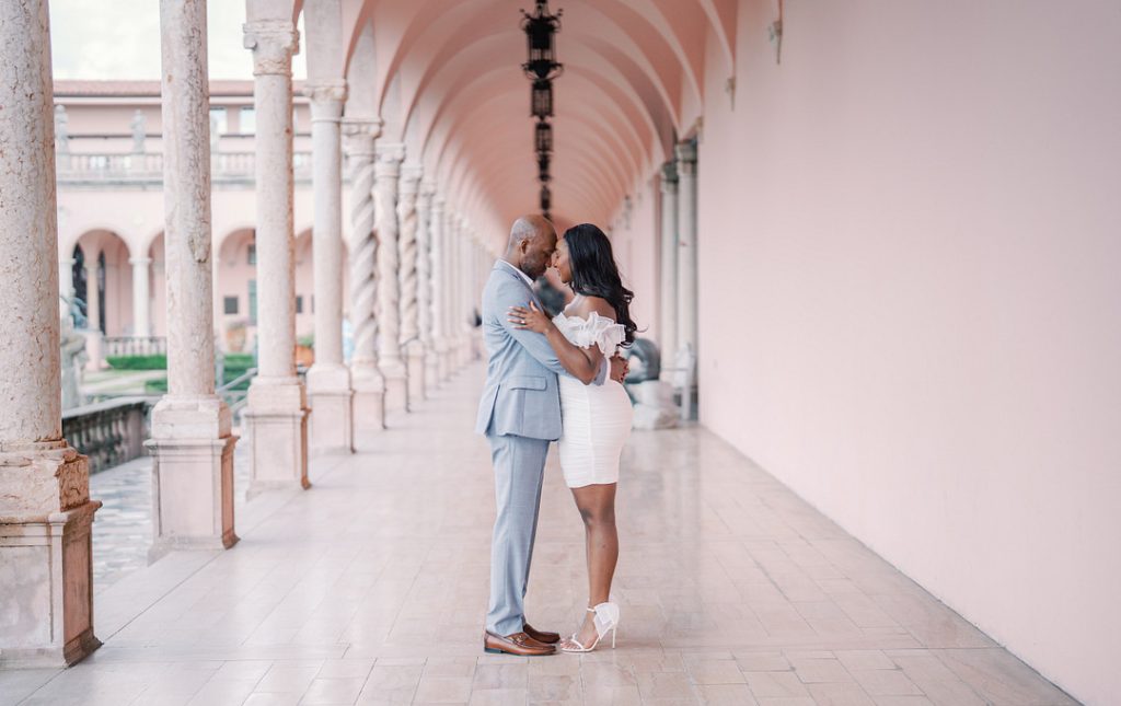 An elegant Black couple embraces affectionately, touching foreheads while standing in a long, pink-walled loggia lined with classical stone columns.