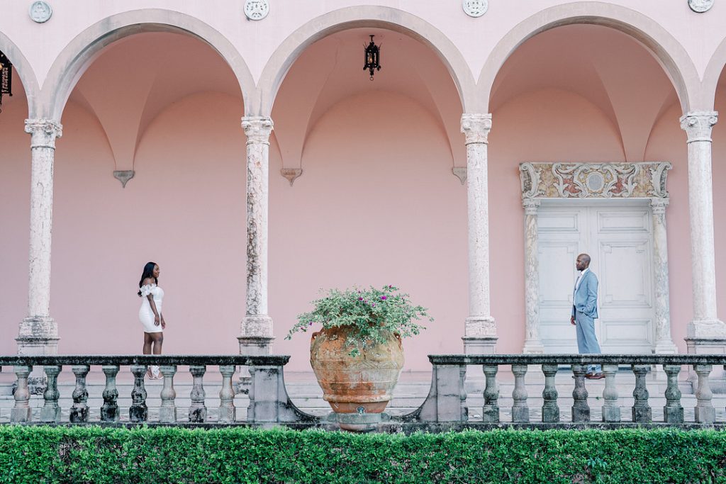 A well-dressed Black couple stands at a distance from each other, looking at one another on a stone balcony with pink arches, separated by a large potted plant.