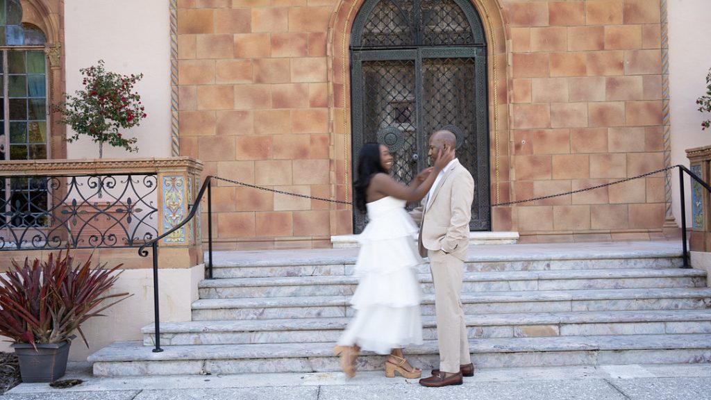 A smiling woman in a tiered white dress gently touches the face of a man in a beige suit as they stand together on stone steps in front of an ornate, arched doorway.