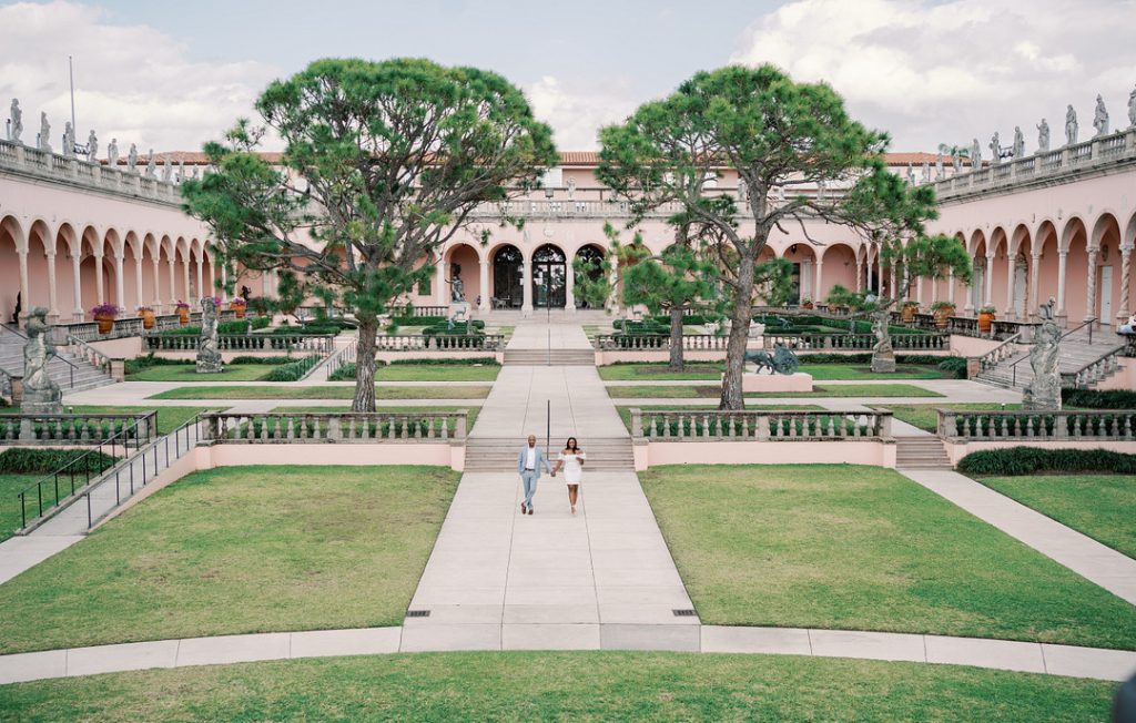 A couple holds hands while walking down a central stone pathway in a grand, symmetrical courtyard featuring manicured lawns, large pine trees, and a pink, arched Italianate building adorned with statues.