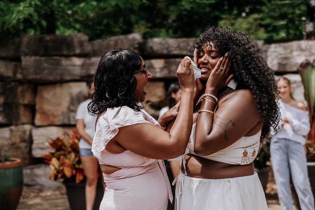 An older woman in a pink dress lovingly wipes tears from the face of a smiling, emotional younger woman with curly hair during an outdoor gathering.