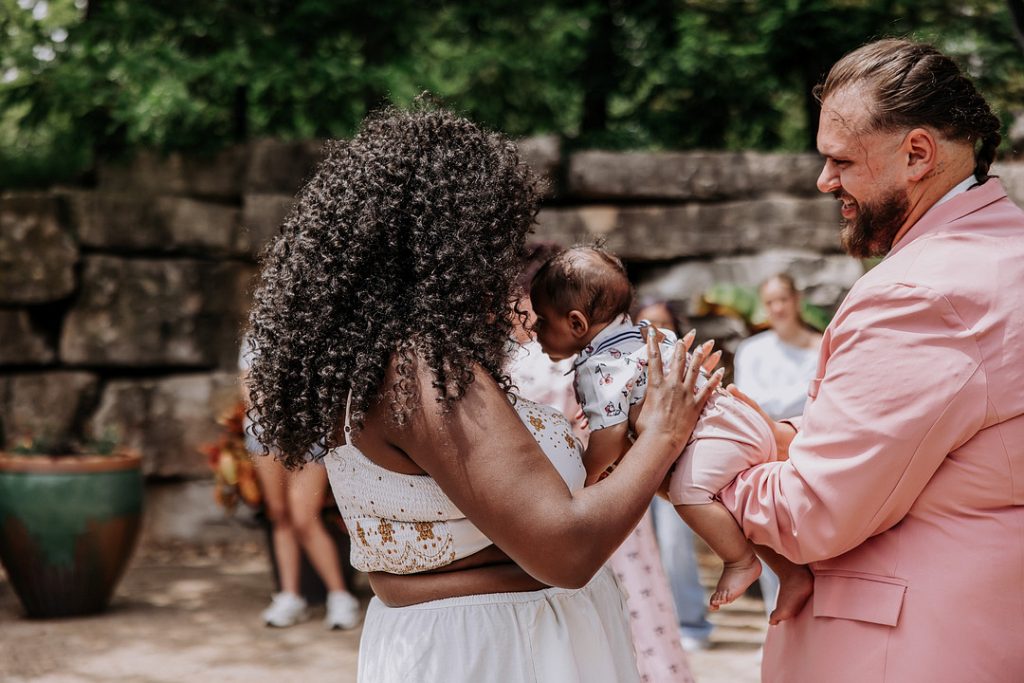 A smiling man in a pink suit and a woman with voluminous curly hair lovingly hold a baby together outdoors in front of a stone wall.