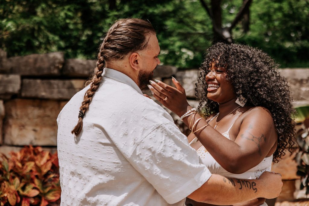 A smiling Black woman with curly hair lovingly touches the face of a man with a braided ponytail as they embrace outdoors.