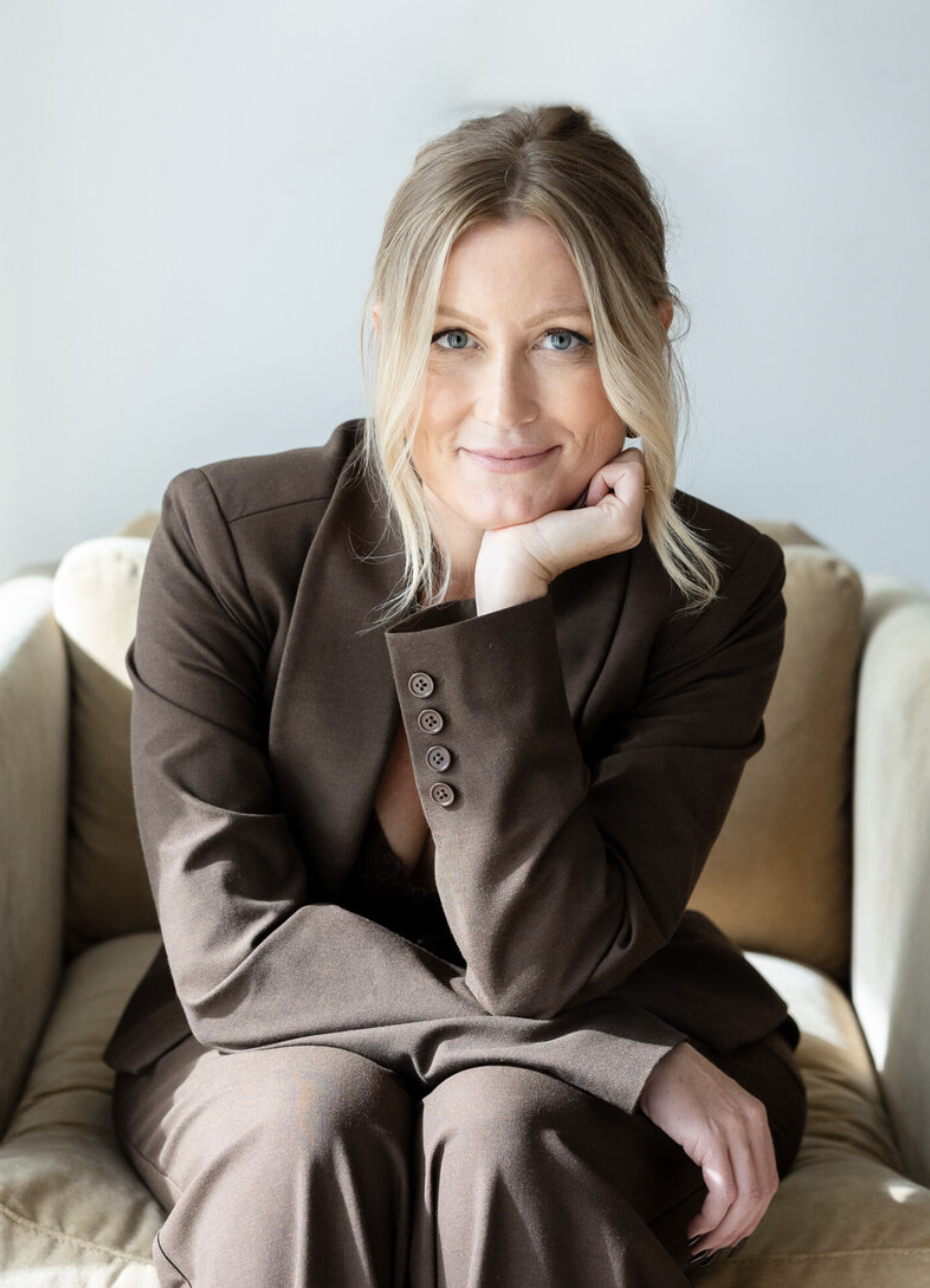 A blonde woman with blue eyes sits in a tan chair, wearing a brown suit and resting her chin on her hand while looking at the camera. The suit jacket has four buttons, and her hair is pulled back with strands framing her face.