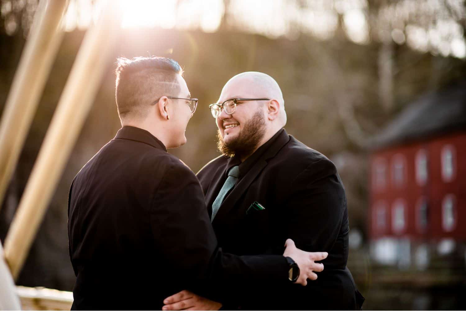 Two husbands hold one another and smile as sunlight spills onto them from behind
