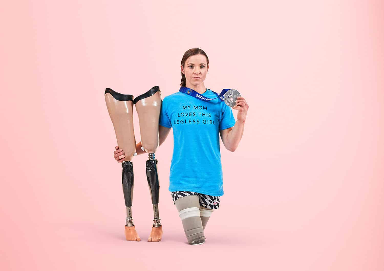 Paralympic gold medalist Oksana Masters stands beside her prosthetics on a pink background showing off her silver medal and wearing a tee-shirt that says