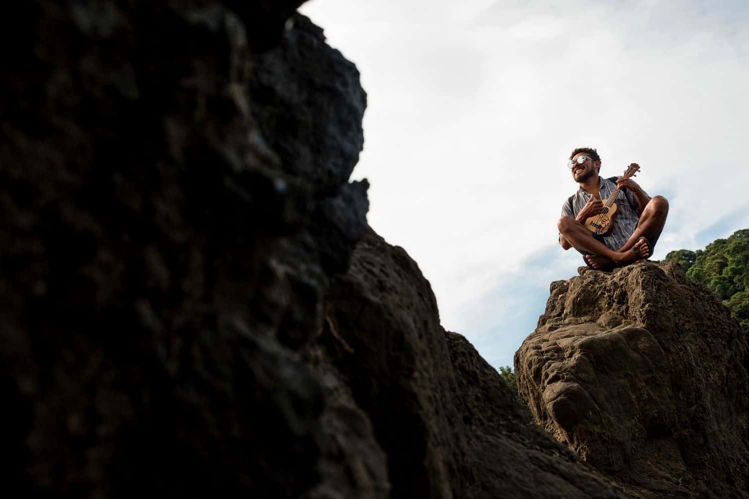A man sitting on top of a large boulder while playing his ukulele