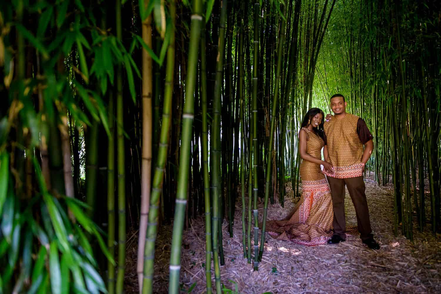 A Black couple poses wearing traditional African attire surrounded by towering bamboo shoots