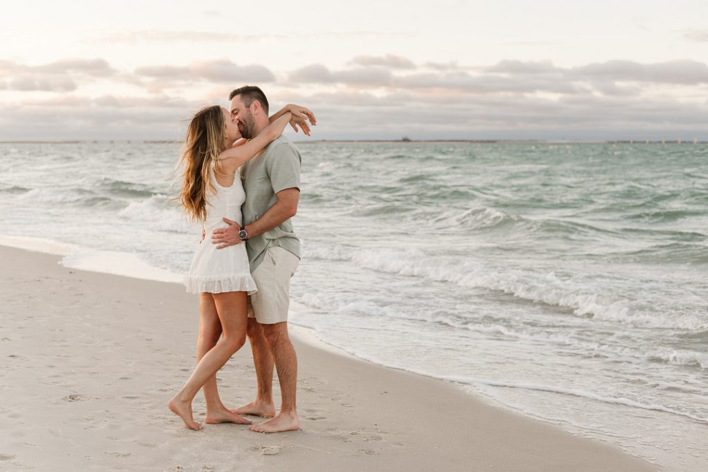 A barefoot couple embraces and kisses on a sandy beach as gentle ocean waves roll in under a cloudy sky.