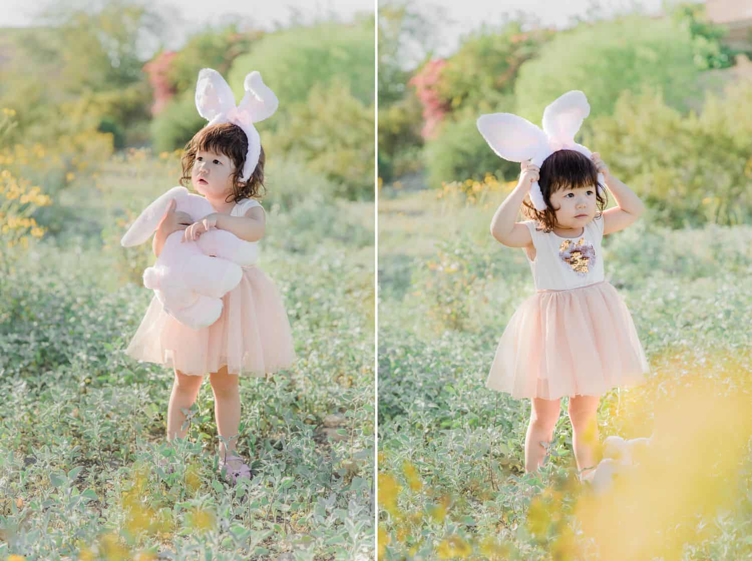 A little girl in a pink dress stands in a field wearing bunny ears