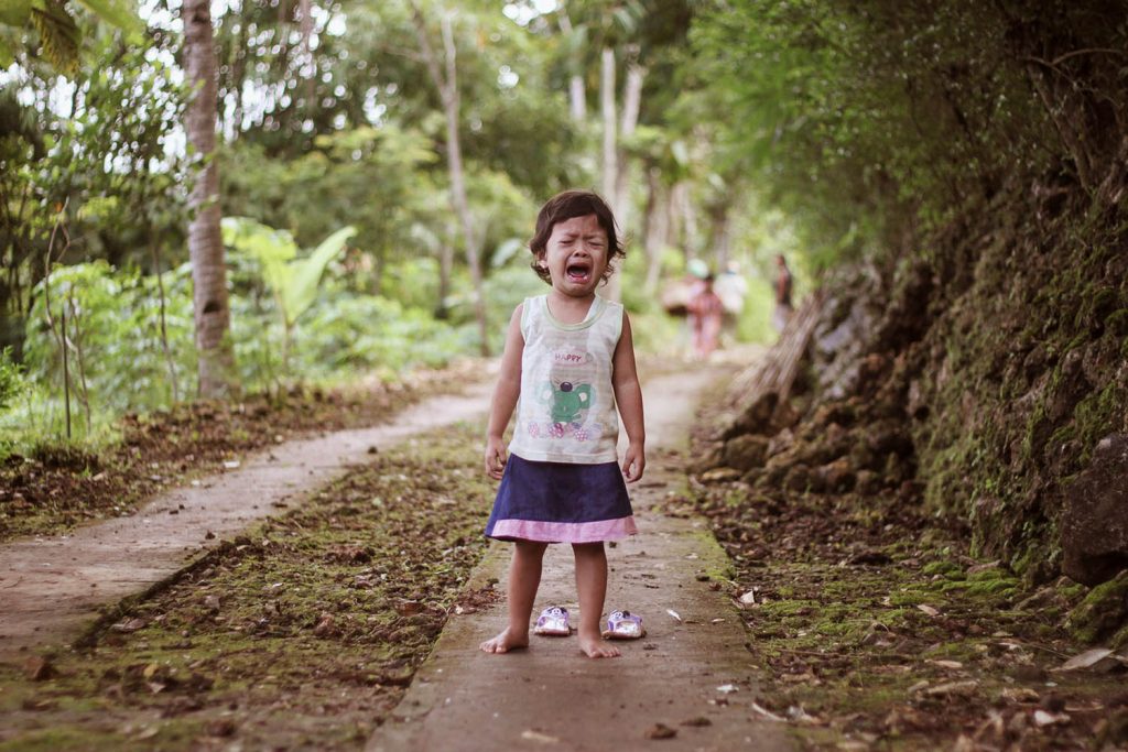 A little girl stands on a sidewalk with her shoes on the ground behind her. She is crying.