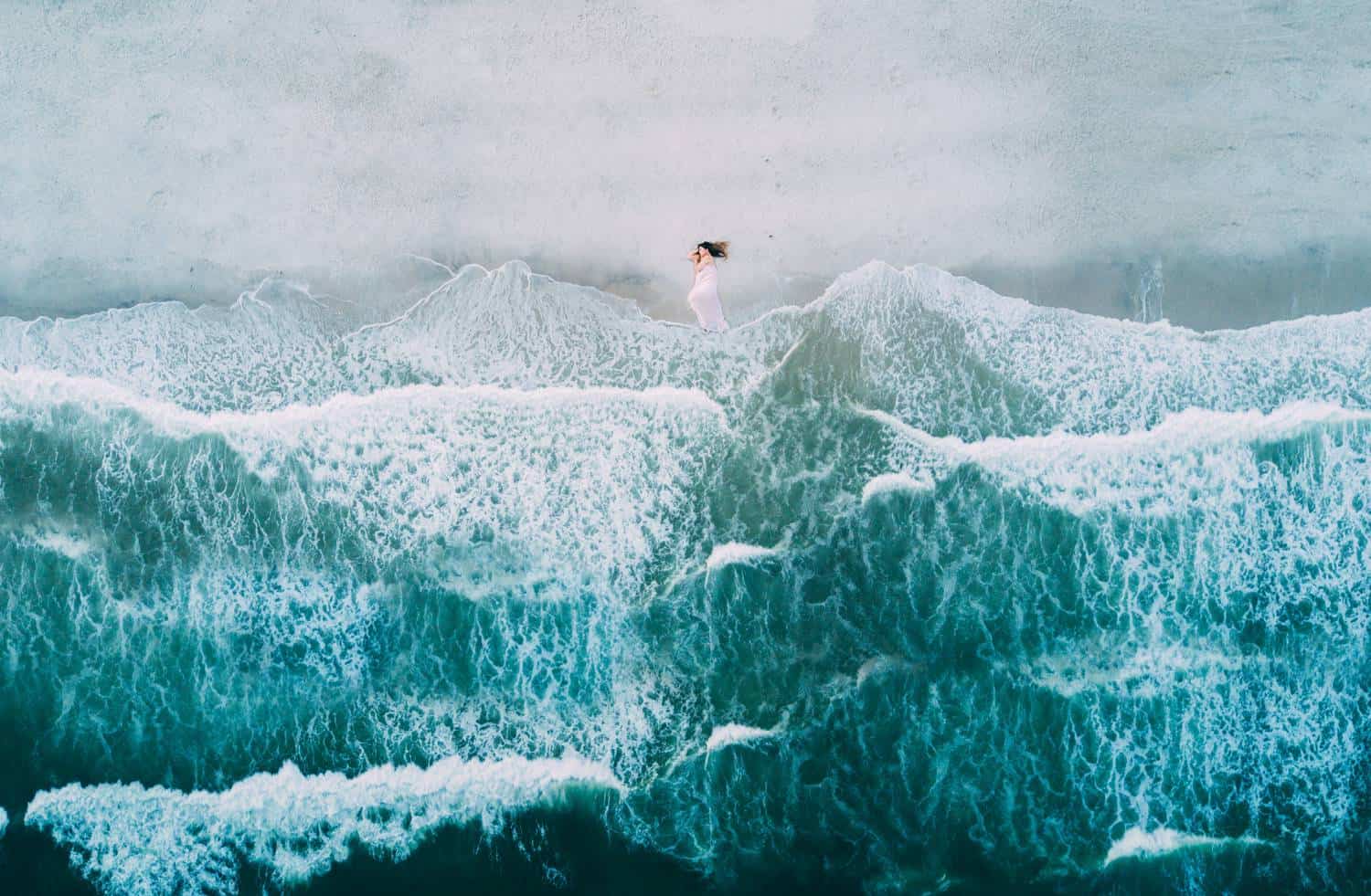 A fine-art drone photograph depicts the tiny image of a white-dressed pregnant woman reclining on a beach above a bright blue ocean's waves.
