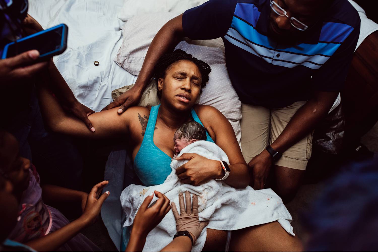 A mother embraces her newborn against her chest after just giving birth. She is surrounded by the supportive arms of her friends and family.