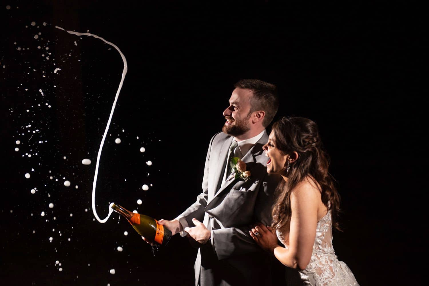 A bride and groom laugh as they pop a bottle of champagne