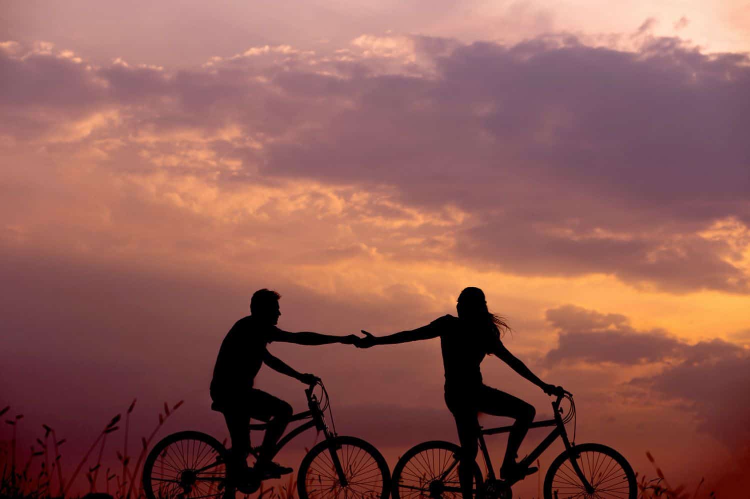 Two silhouetted cyclists hold hands in front of a sunset.