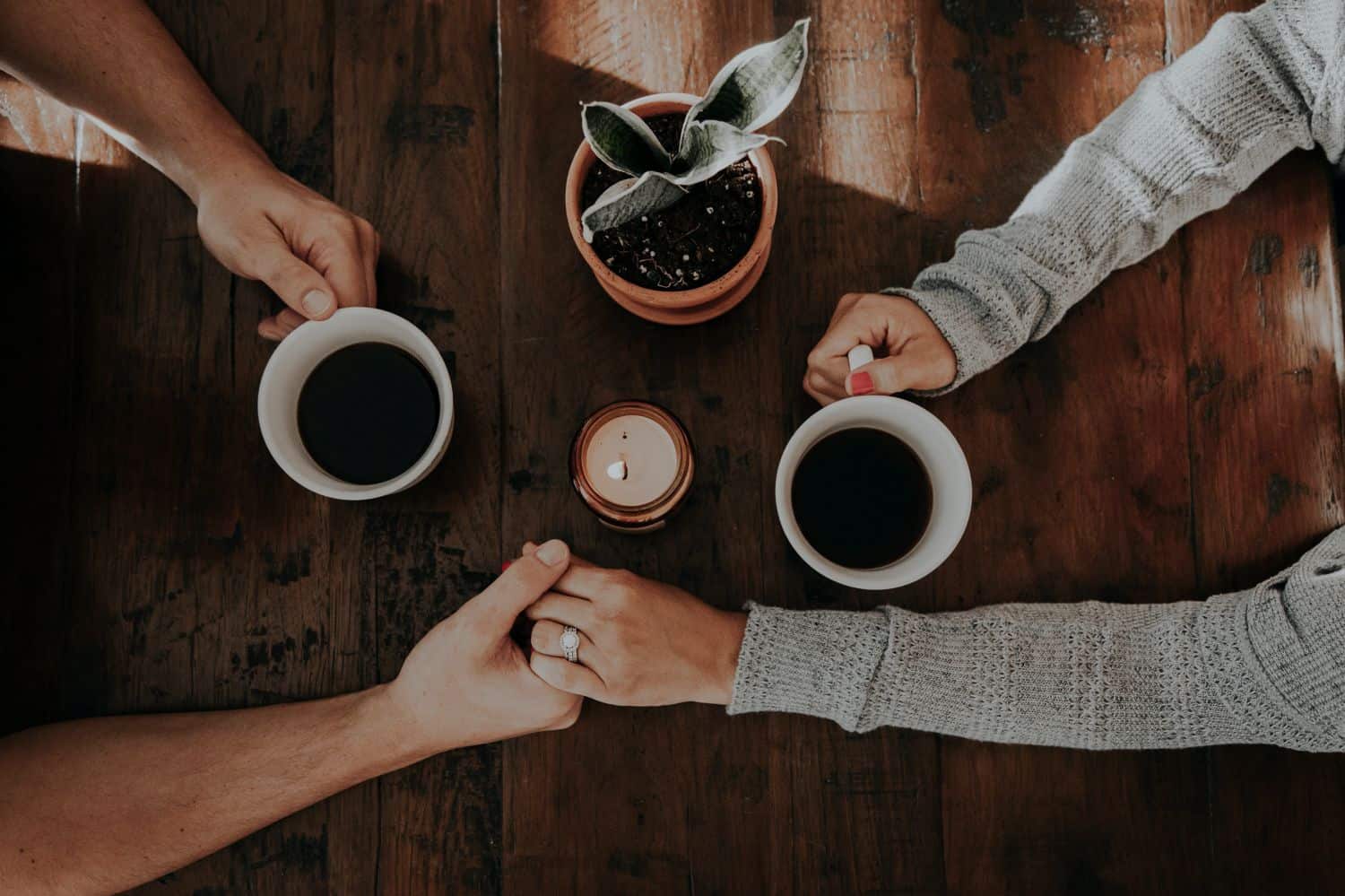 Shown from above, two people hold hands across a wooden table. They each have a cup of coffee and a small plant sits between them.