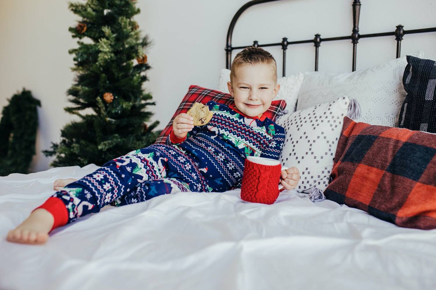 In this photo by Logan Fahey, a little boy wearing Christmas pajamas reclines on a white bed with a cookie and a red mug of hot cocoa in his hands. Christmas Mini Session: Ideas Families will ADORE!