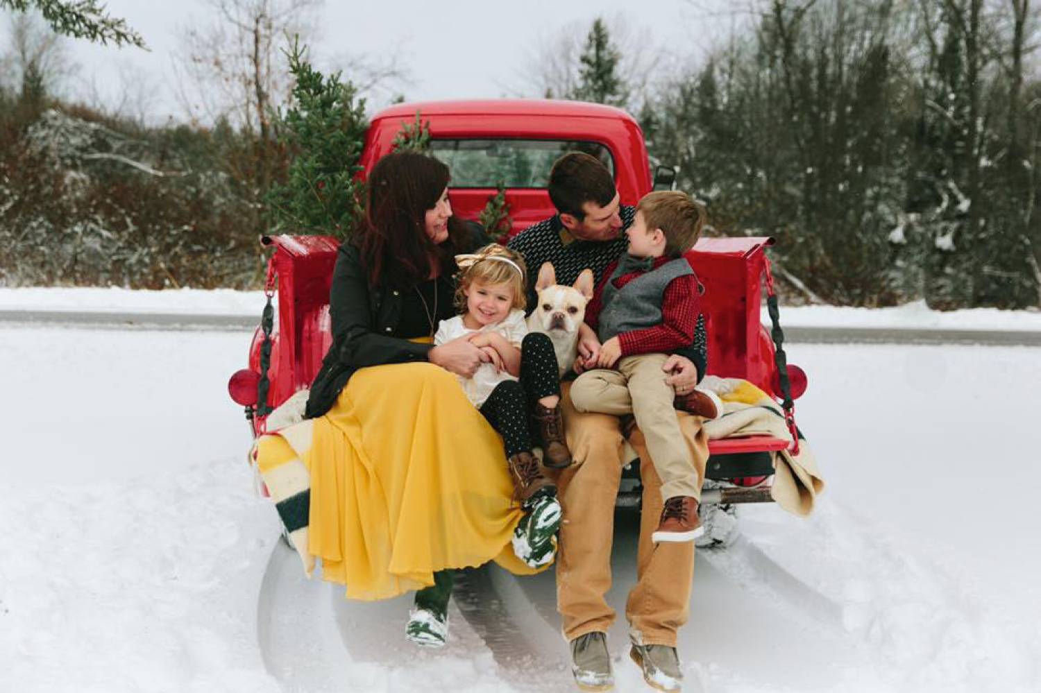 In this photo by Maine Tinker Photography, a mom, dad, two kids, and a small dog sit in the back of a vintage red truck that's parked in the snow. Christmas Mini Session: Ideas Families will ADORE!