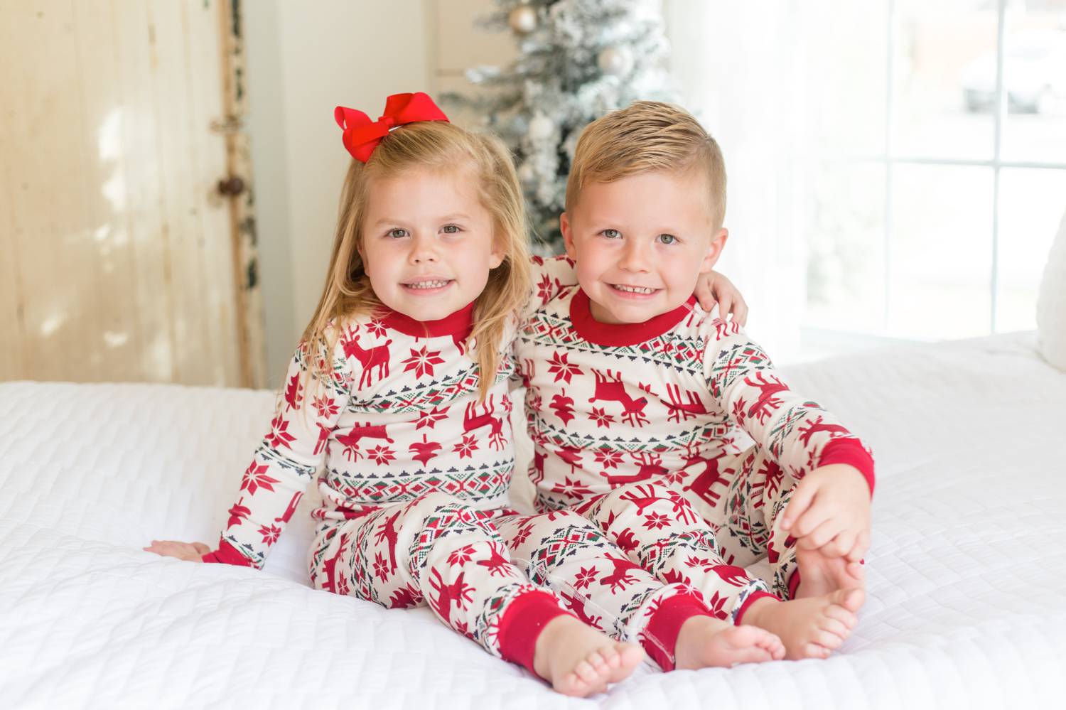 In this photo by Rebecca Rice, toddler twins wearing Christmas pajamas sit on a white-draped bed with their arms around each other as they smile at the camera. Christmas Mini Session: Ideas Families will ADORE!