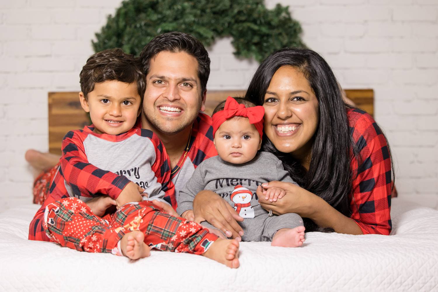In this photo by Rebecca Rice, a family of color is photographed on a white-draped bed in a white brick studio. The parents are lying on their stomachs, facing the camera and smiling. Their young son and baby daughter sit on the bed with their parents' arms wrapped around them. A Christmas wreath hangs on the wall behind them. Christmas Mini Session: Ideas Families will ADORE!