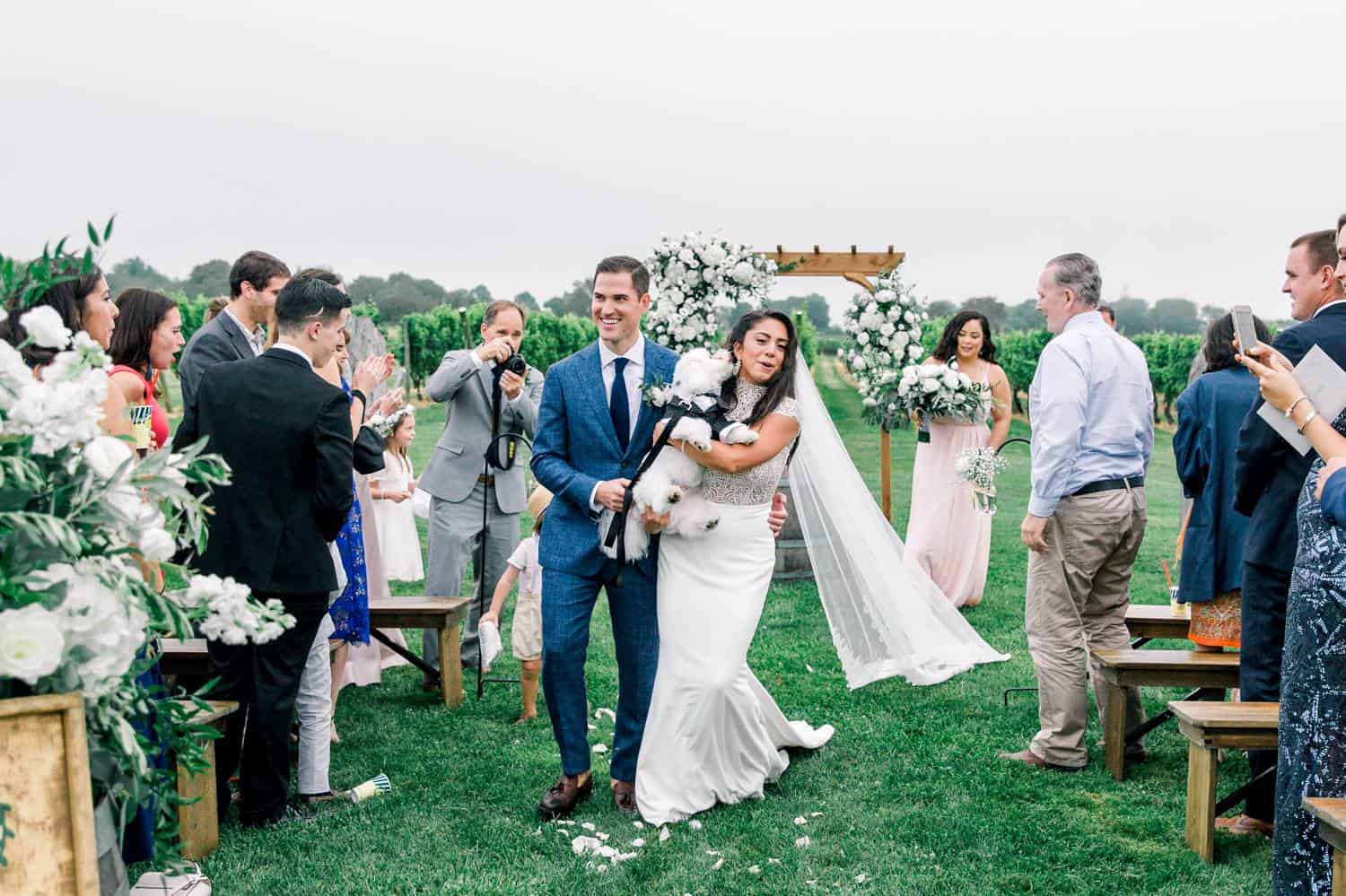 A newly married couple walks down the aisle carrying their fluffy white dog as their guests stand and cheer.