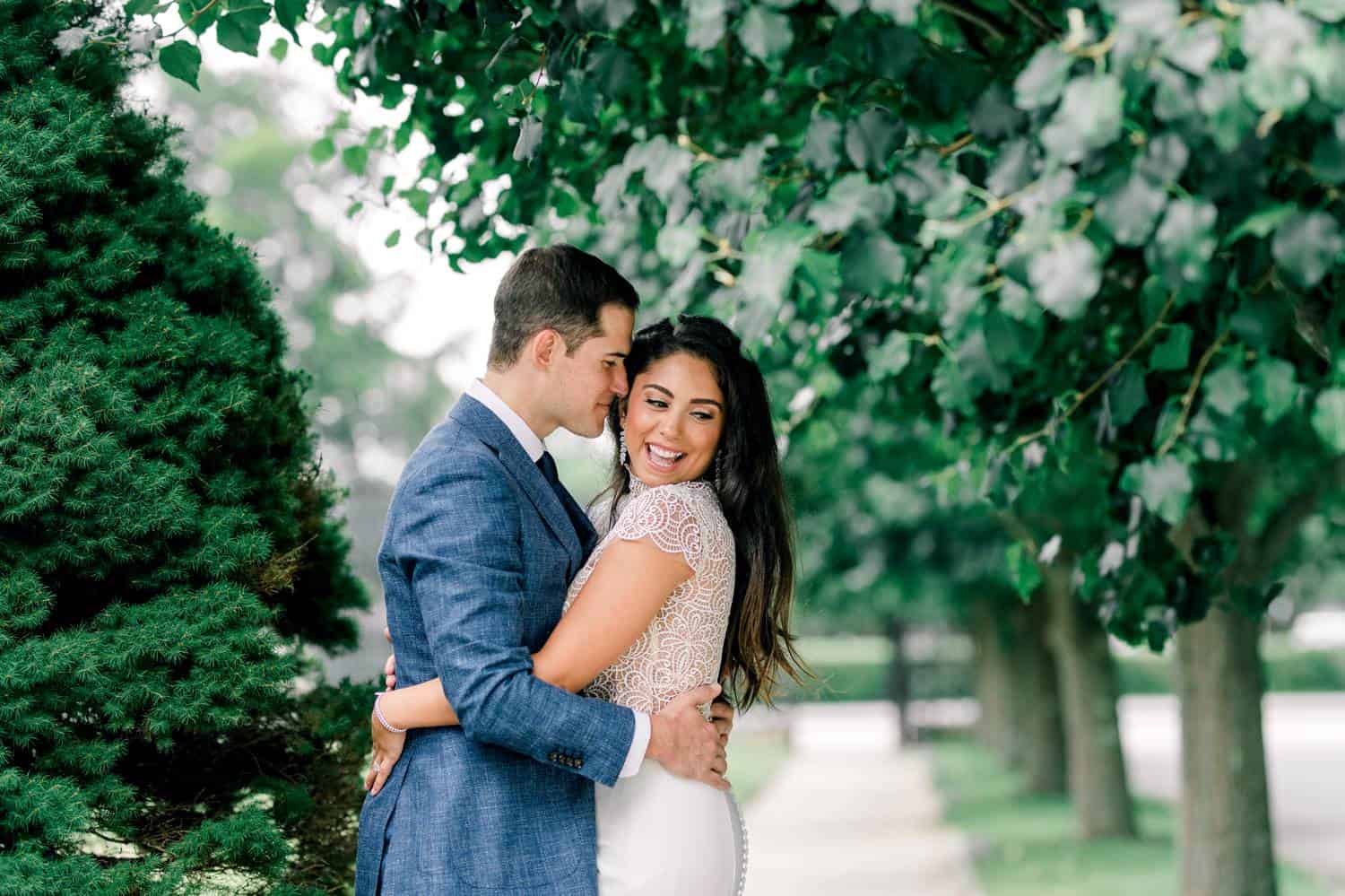 A bride and groom stand outside on a shady sidewalk. The groom embraces the bride with his face pressed against the side of her face.