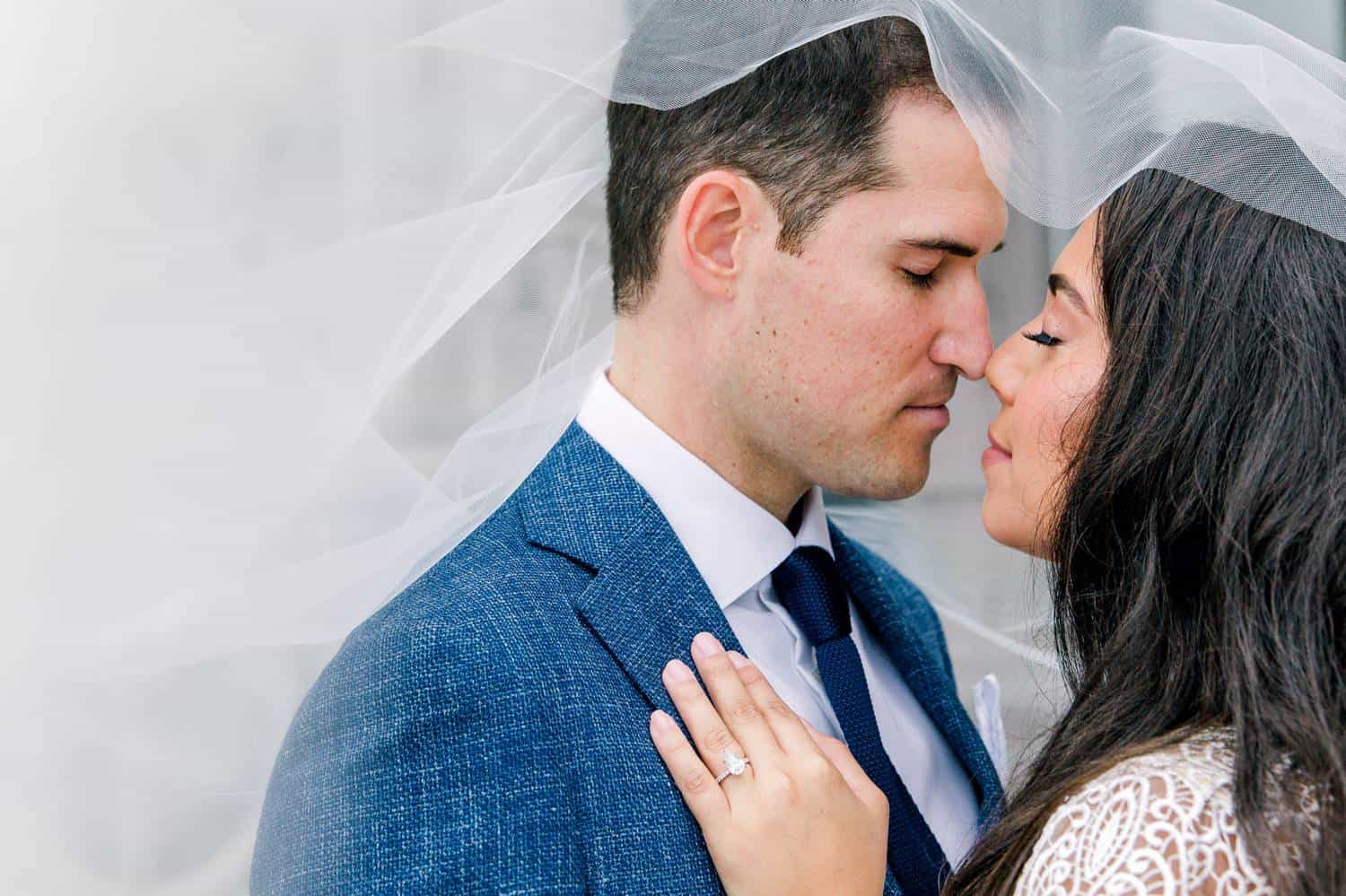 A bride and groom stand nose-to-nose with their eye closed. The bride's veil is draped over their heads and puffed out around them like a cloud.