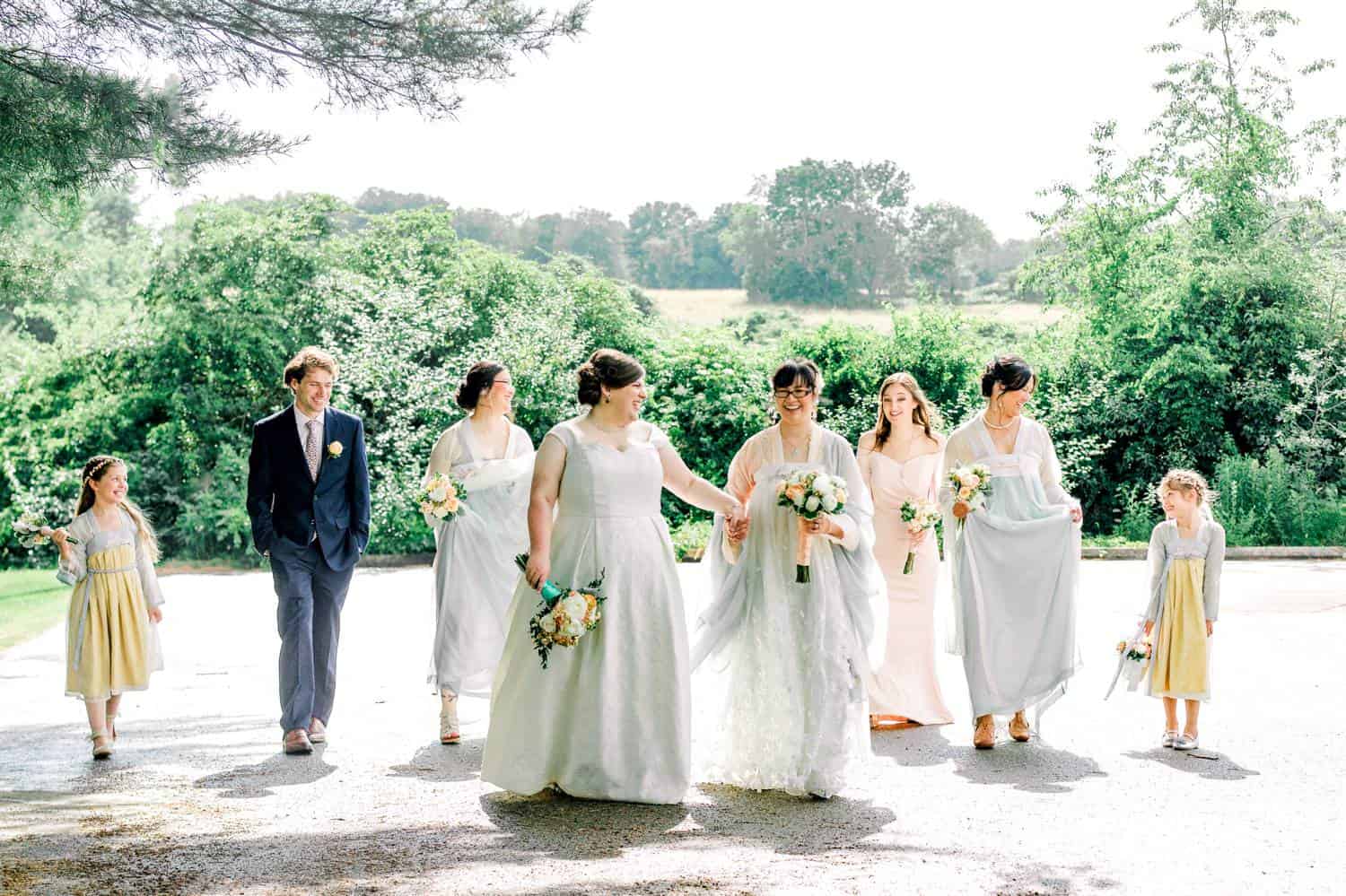 Two brides stroll under a tree. They are backlit by the bright sun, and surrounded by their wedding party.